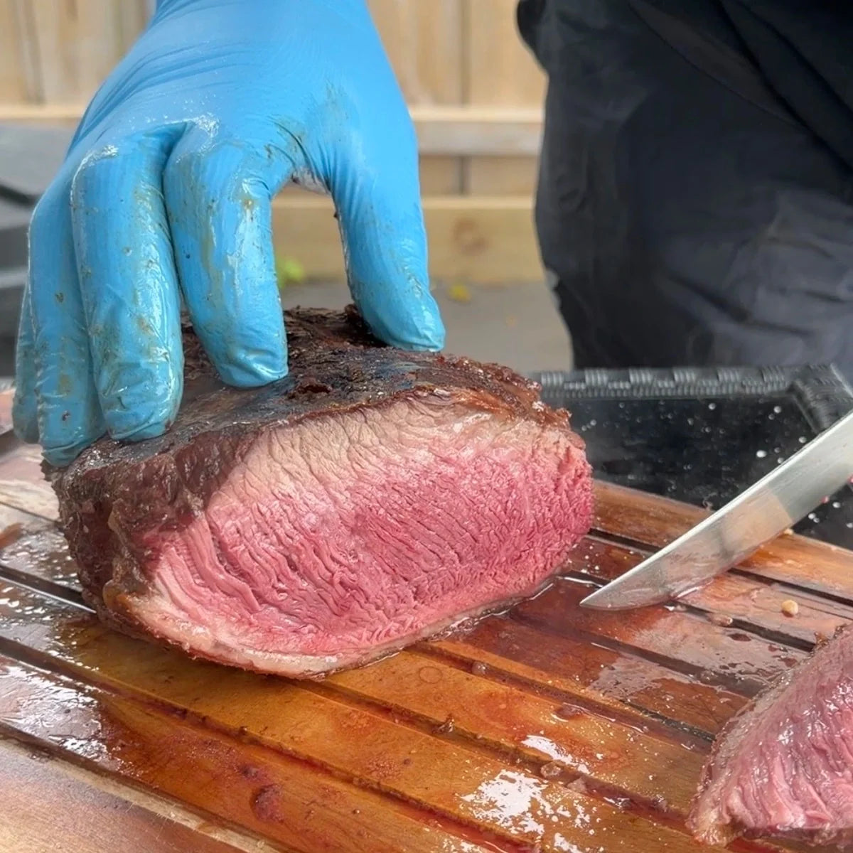 Whole picanha (rump cap) with a dark crust being sliced on a wooden board, showing an even pink centre