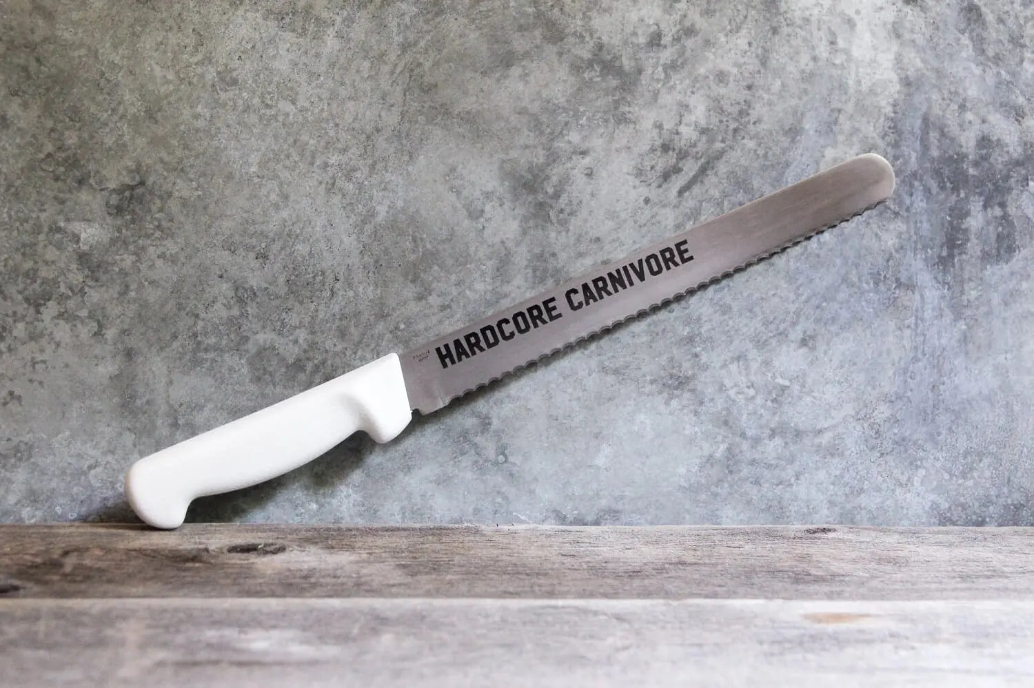 Hardcore Carnivore serrated brisket slicing knife with stainless steel blade and white ergonomic handle on rustic surface.