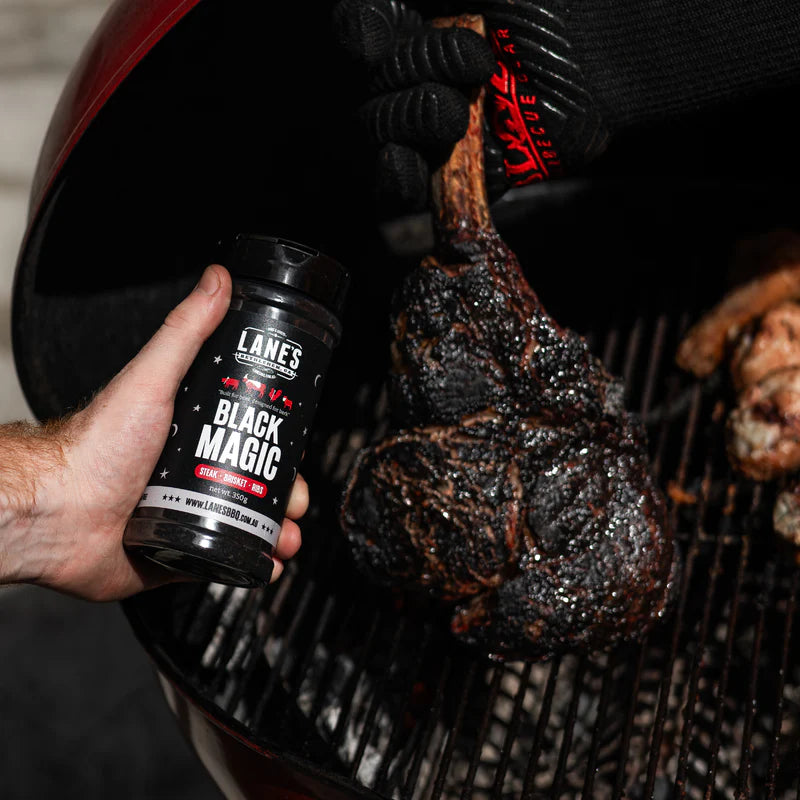 Tomahawk steak cooking on BBQ alongside Lane’s Black Magic seasoning bottle