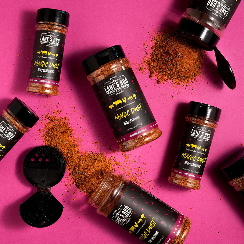Lane’s Magic Dust BBQ rub bottles displayed with seasoning scattered on a pink background