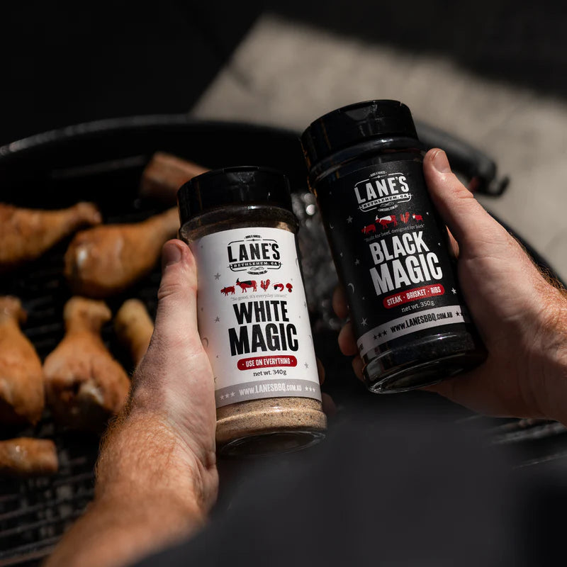 Hands holding Lane’s White Magic and Black Magic rubs over BBQ grill with chicken