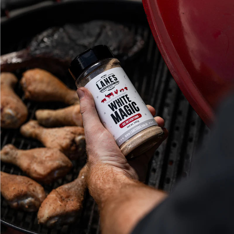 Lane’s White Magic BBQ rub held over a grill with chicken drumsticks cooking