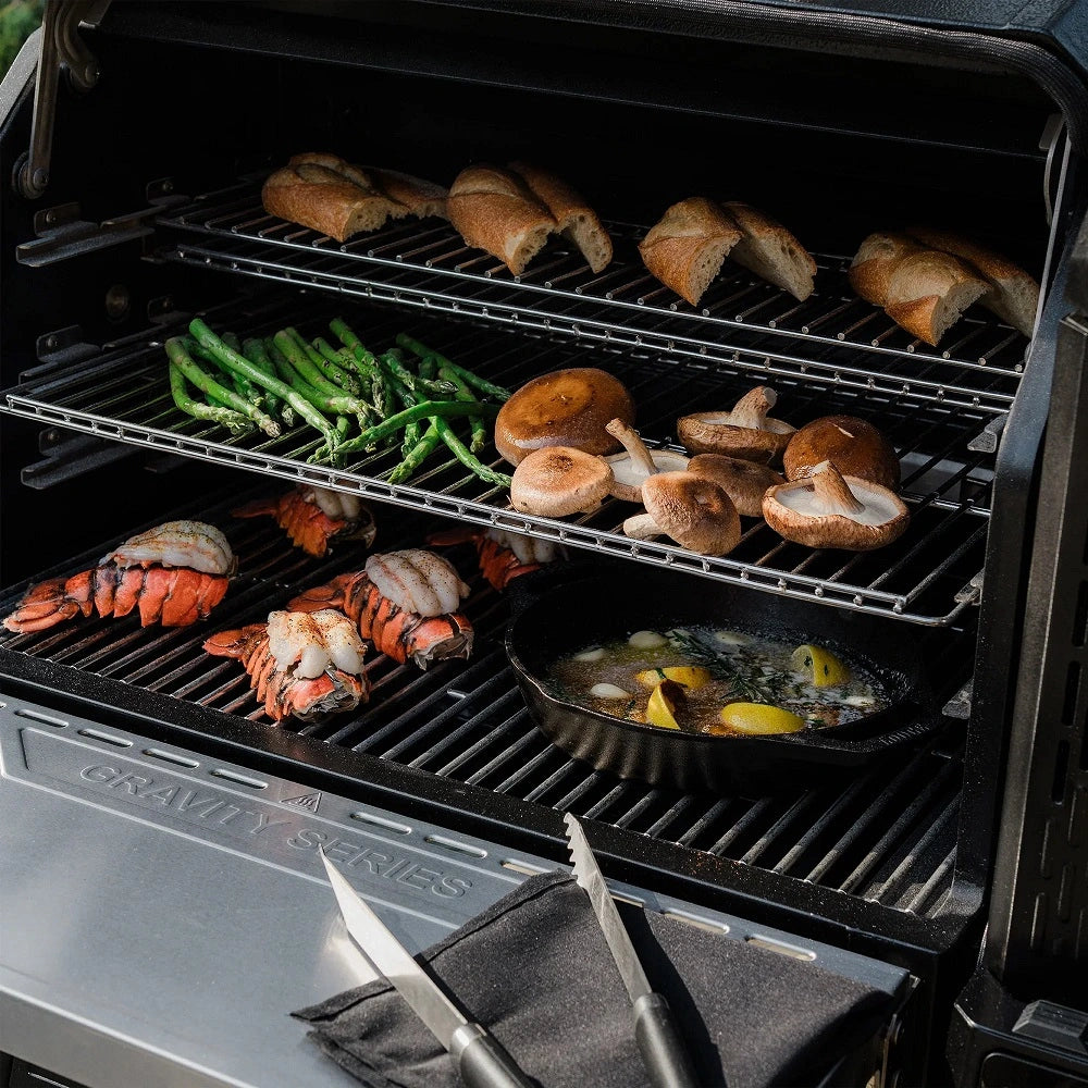 Food cooking inside the Masterbuilt Gravity Series XT Digital Charcoal BBQ showing multiple cooking racks with seafood, vegetables, mushrooms and bread.
