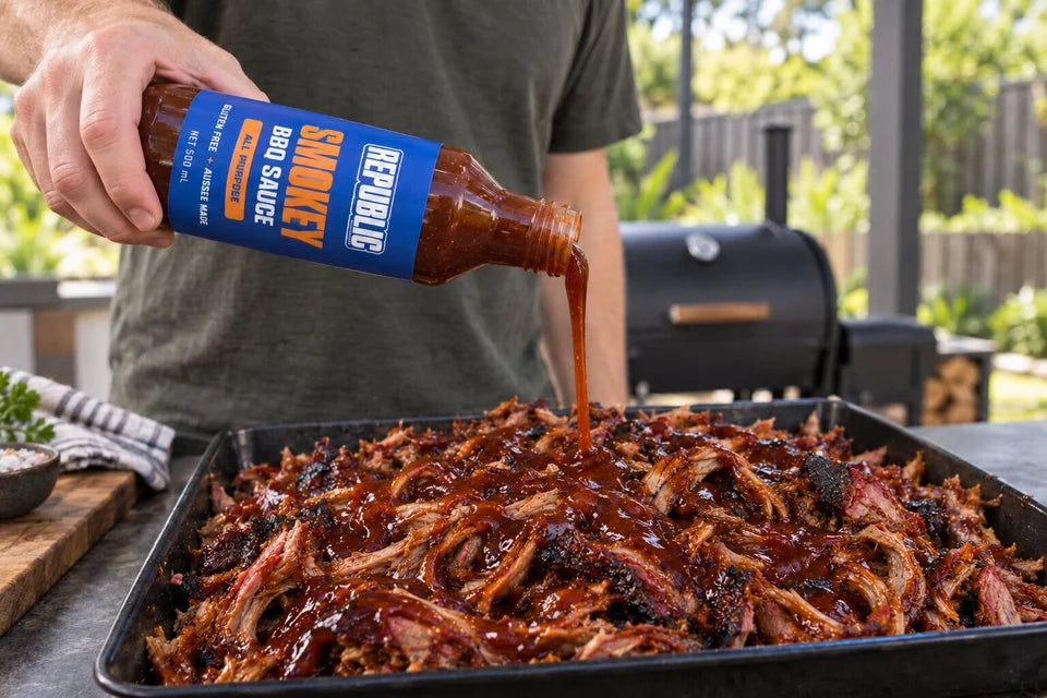 Republic Smokey BBQ Sauce being poured over a tray of pulled pork beside a barbecue